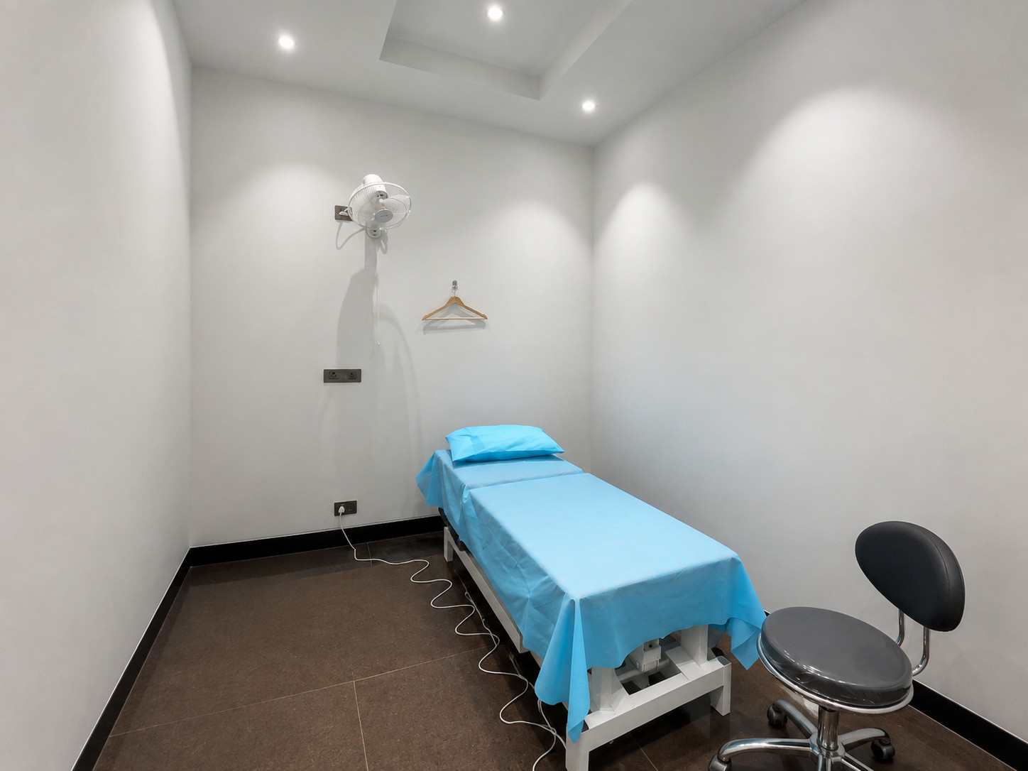 Physiotherapy Treatment Room with Therapy Bed – Clinic Interior by Nail & Hammer Private physiotherapy treatment room with an adjustable therapy bed draped in blue linen, a wall-mounted fan and a stainless steel stool in a clinic interior designed by Nail & Hammer